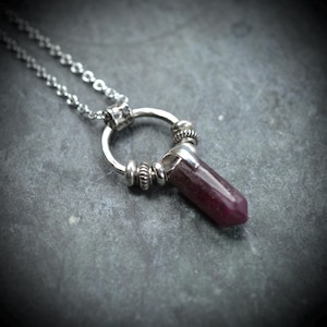 May include: A silver-toned necklace featuring a polished, deep red, hexagonal crystal pendant suspended from a circular metal ring. The ring is accented with small, textured silver beads and connects to a delicate silver chain with round links.