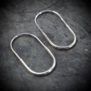 May include: A pair of silver-coloured hoop earrings with an elongated oval shape. Each earring features a continuous wire design with a smooth, polished metallic finish and a simple closure mechanism where the wire ends meet to form a seamless loop.