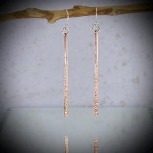 May include: A pair of copper-coloured, textured bar earrings with silver-coloured ear wires. The earrings are suspended from a small, brown branch against a light background. The earrings have a vertical, linear design.