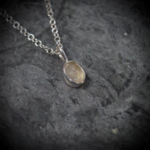 May include: A silver necklace featuring a small, oval-shaped, translucent pale yellow gemstone pendant set in a smooth silver bezel. The pendant hangs from a delicate silver chain with round links, creating a simple and refined jewellery piece.