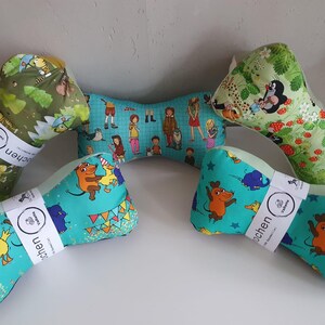Könnte beinhalten: Fünf knochenförmige Stoffkissen mit verschiedenen bunten Cartoon-Drucken. Drei Kissen sind mit weißen Papierbändern umwickelt, die mit ValHa beschriftet sind. Die Muster zeigen grüne Waldszenen, ein blaues Gitter mit Figuren und helles Türkis mit Tieren.