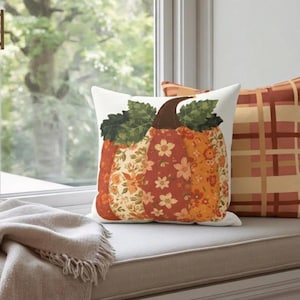 May include: Two square throw pillows. One pillow features a large pumpkin graphic composed of various floral patterns in shades of orange, red, and yellow with green leaves. The second pillow has a plaid pattern in orange, brown, and cream tones.