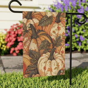 May include: A rectangular garden flag hanging on a black metal stand, featuring a repeating pattern of pumpkins in shades of orange, cream, and brown, accented with dark green leaves and small floral motifs across the entire surface.