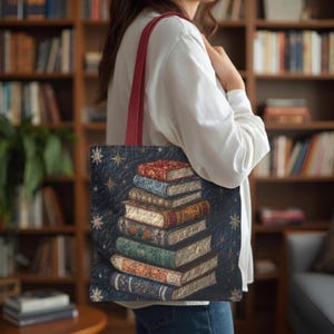 May include: A navy blue tote bag with a stack of books on the front. The books have colorful covers and are decorated with floral and geometric patterns. The bag has a red strap and is carried over the shoulder.