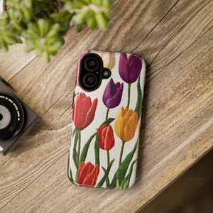 May include: A white phone case with a floral design featuring colourful tulips in shades of red, yellow, purple, and orange. The case has a camera cutout and a white background.