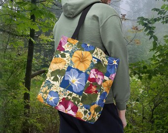 May include: A floral tote bag with a patchwork design featuring blue, yellow, and red flowers. The bag has black straps and is being held by a person.