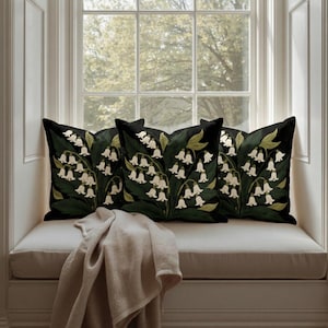 May include: Three black throw pillows with a white lily of the valley floral design. The pillows are arranged on a window seat with a beige blanket. The window has a view of trees.
