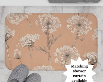 May include: A peach-colored bath mat with a white floral pattern. The mat features a repeating design of delicate white flowers with thin black stems. The text "Matching shower curtain available" is in a white starburst shape at the bottom right corner of the mat.