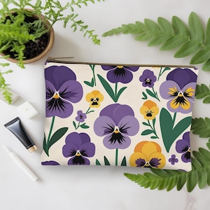 May include: A rectangular beige zippered pouch featuring a repeating pattern of purple and yellow pansies with green leaves. The flowers have black centers. The pouch is lying on a white surface with greenery.