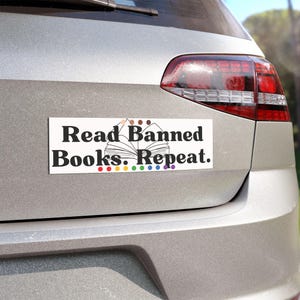 May include: A rectangular white bumper sticker with the text "Read Banned Books. Repeat." in black. The design includes an open book graphic and colorful dots. The sticker is affixed to the rear of a silver car.