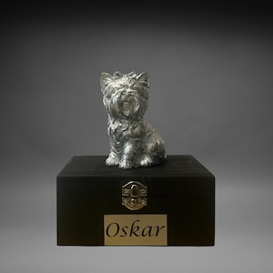 May include: A silver-colored Yorkshire Terrier figurine sits atop a black wooden box. The box has a gold-colored nameplate that reads "Oskar". The figurine is detailed with textured fur and is in a seated position. The box has a gold-colored clasp.