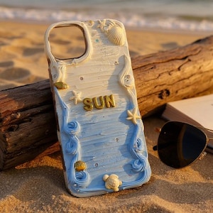 Puede incluir: Funda de teléfono blanca y azul con temática de playa, con la palabra "SUN" en letras doradas, conchas marinas y estrellas de mar. La funda está sobre arena cerca de un tronco, un libro abierto y gafas de sol.