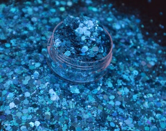 May include: A small glass jar filled with teal blue glitter. The glitter is scattered around the jar on a black surface. The number 37 is written on a white square in the upper left corner of the image.