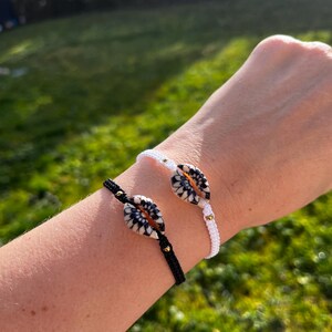 Peut inclure: Deux bracelets faits à la main, un noir et un blanc, chacun avec une pièce maîtresse en forme de coquillage décoratif. Les coquillages ont un motif noir et blanc avec des accents orange. Des perles dorées sont utilisées comme ornements sur les bracelets.