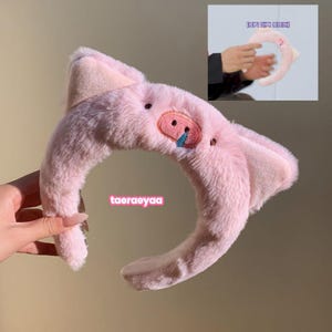 Puede incluir: Una diadema rosa de felpa con orejas de cerdo y un diseño de cara de cerdo. La cara del cerdo tiene una boca roja y una lágrima azul. El texto "taeraeyaa" está en la diadema.