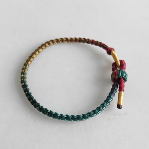 Op de afbeelding: Een gevlochten armband met een groene, gouden en rode kleurstelling. De armband is aan het einde met een knoop vastgemaakt.