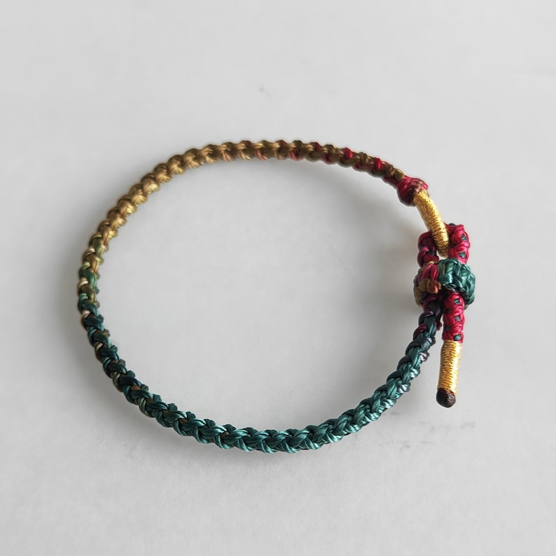 Op de afbeelding: Een gevlochten armband met een groene, gouden en rode kleurstelling. De armband is aan het einde met een knoop vastgemaakt.