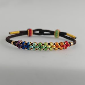 Peut inclure: Un bracelet de couleur arc-en-ciel avec de petits nœuds en forme de fleur. Le bracelet est fait d'un cordon noir avec des accents dorés et deux petites perles dorées.