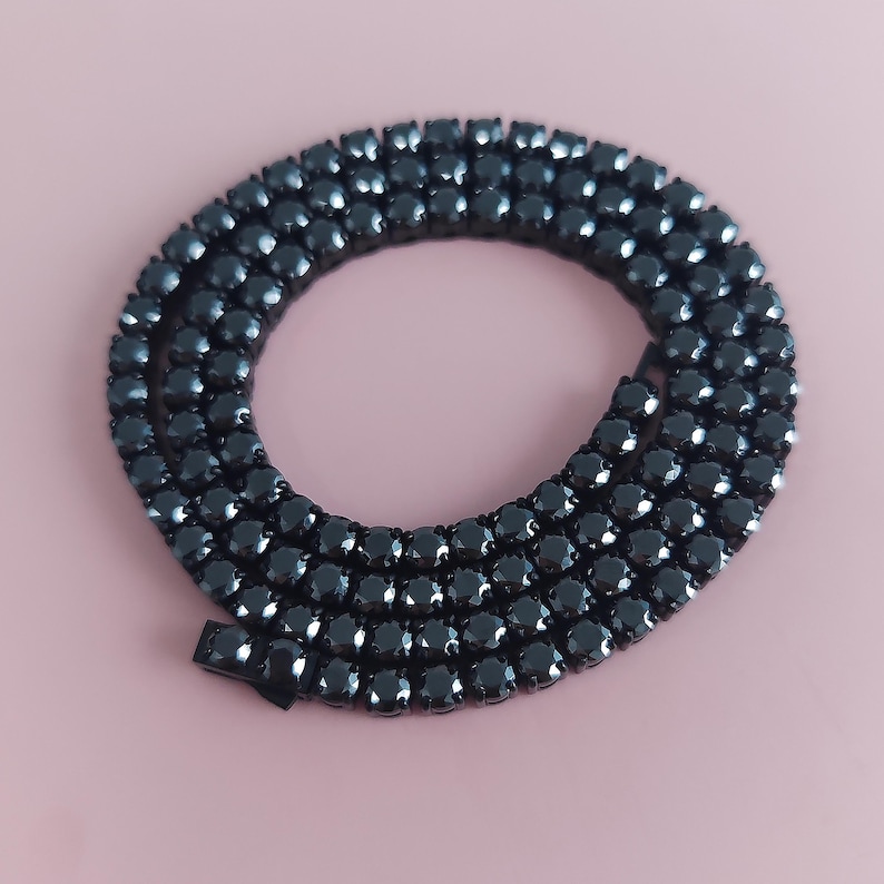 May include: A black diamond tennis chain necklace with a clasp closure. The chain is made of small, round black diamonds that are set close together.