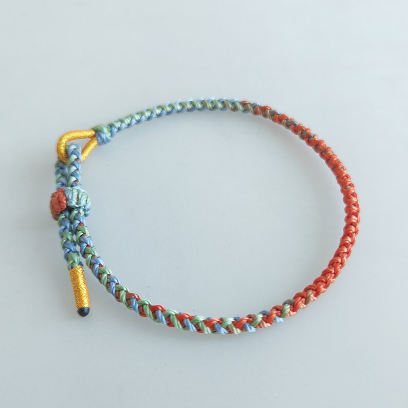Op de afbeelding: Een gevlochten armband met een mix van blauwe, groene en oranje draden. De armband heeft een gouden sluiting.