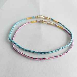 Peut inclure: Deux bracelets en cordon tressé avec des fermoirs dorés. Un bracelet est turquoise, bleu et vert, et l'autre est rose, violet et argent.