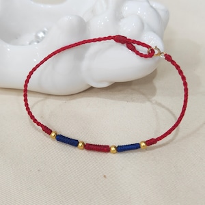 Puede incluir: Un brazalete de cuerda roja con cuentas doradas y detalles azules. El brazalete está atado con un nudo y tiene un cierre.