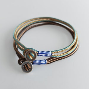 May include: Two braided bracelets with a silver button closure. The bracelets are made of brown, gold, and teal colored cords.