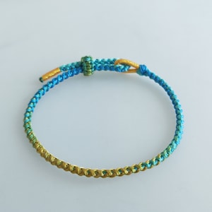 Op de afbeelding: Een gevlochten armband met een gradi&euml;nt van blauwe, groene en gouden kleuren. De armband is gemaakt van een dun, geweven koord en heeft een eenvoudige knoop sluiting.
