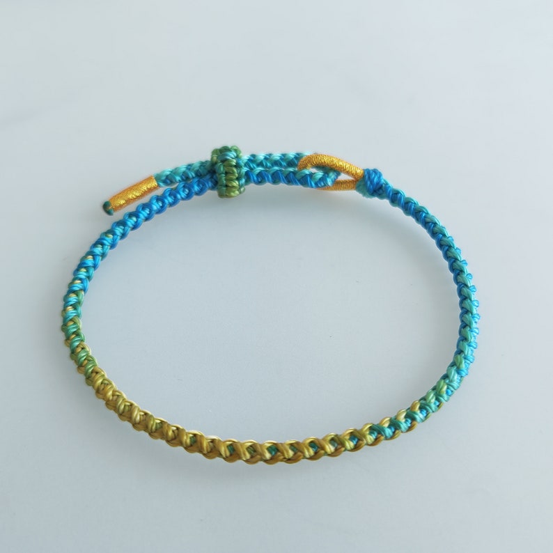 Op de afbeelding: Een gevlochten armband met een gradi&euml;nt van blauwe, groene en gouden kleuren. De armband is gemaakt van een dun, geweven koord en heeft een eenvoudige knoop sluiting.