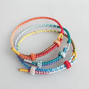 Puede incluir: Tres pulseras trenzadas de colores con detalles anudados. Las pulseras están hechas de hilo rojo, naranja, amarillo, verde, azul y blanco.