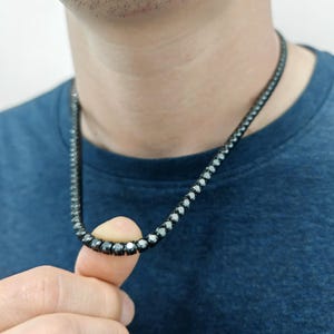 May include: A black diamond tennis chain necklace. The chain is made of small, round black diamonds that are set close together. The necklace is worn by a person, but the person's face is not visible.