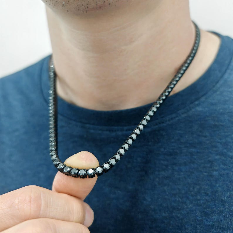 May include: A black diamond tennis chain necklace. The chain is made of small, round black diamonds that are set close together. The necklace is worn by a person, but the person's face is not visible.