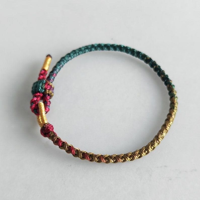 Op de afbeelding: Een gevlochten armband met een knoop aan het einde. De armband is gemaakt van groene, gouden en rode draad.