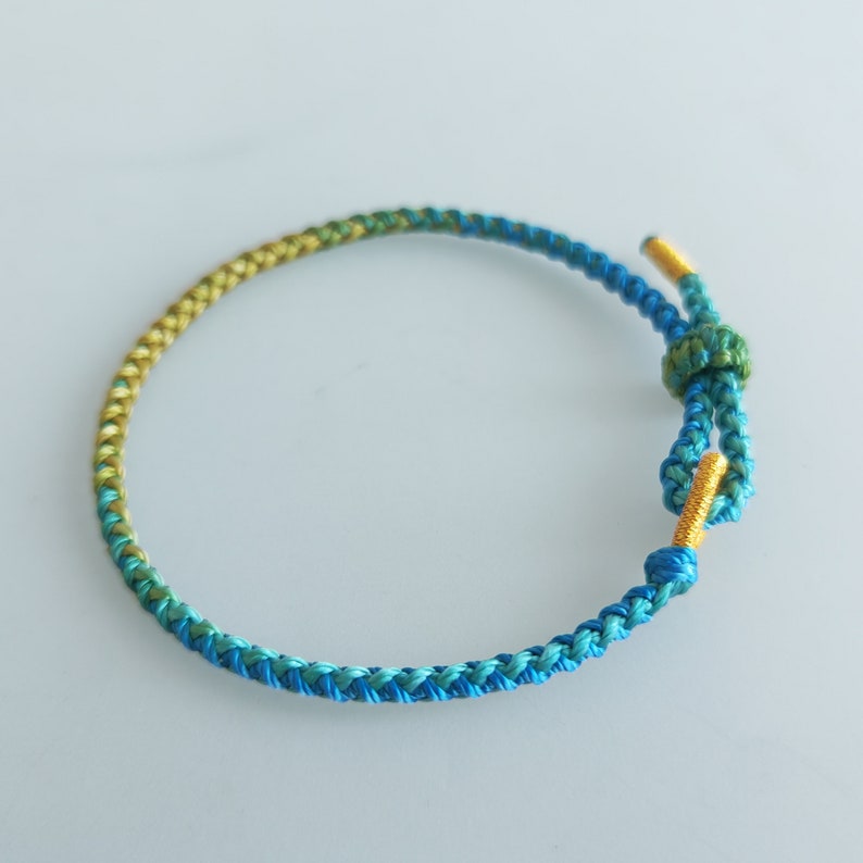 Op de afbeelding: Een gevlochten armband met een gradi&euml;nt van blauwe, groene en gele kleuren. De armband heeft een gouden sluiting.