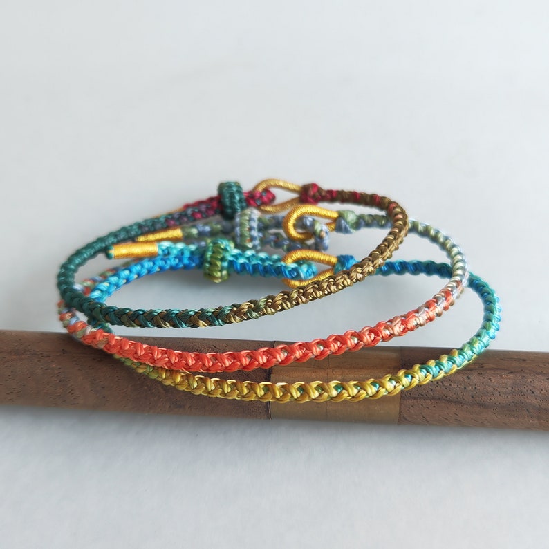 Op de afbeelding: Vier kleurrijke gevlochten armbanden in tinten rood, oranje, geel, groen, blauw en bruin. De armbanden liggen op elkaar gestapeld op een houten oppervlak.