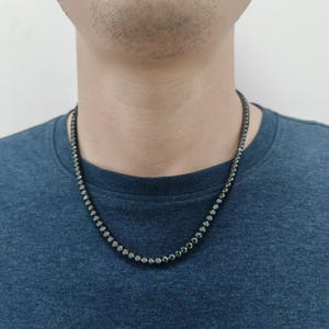 May include: A black chain necklace with small, round, black stones. The necklace is worn by a person with a dark blue shirt.