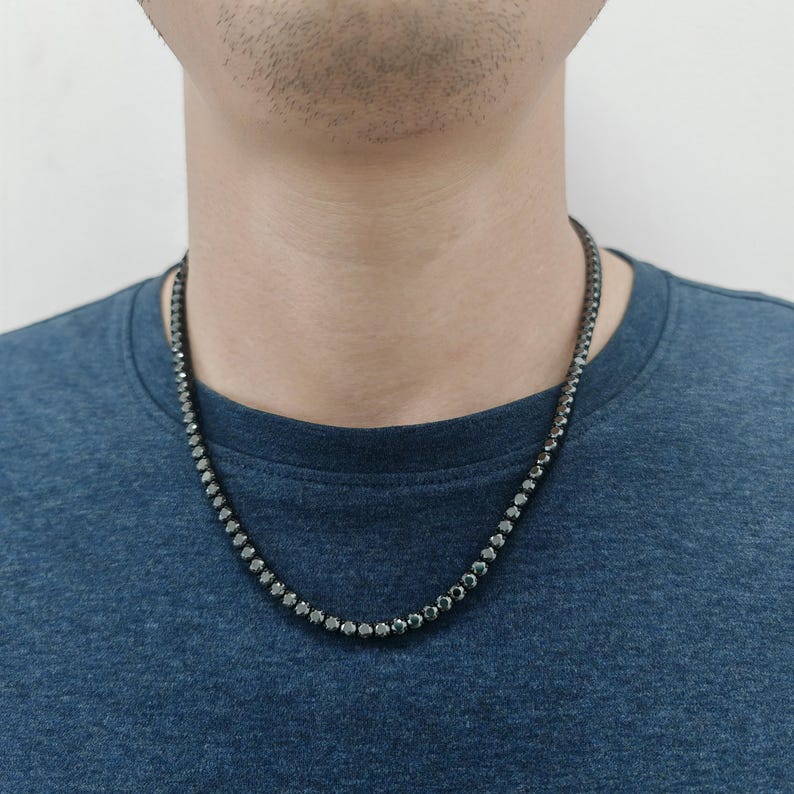 May include: A black chain necklace with small, round, black stones. The necklace is worn by a person with a dark blue shirt.