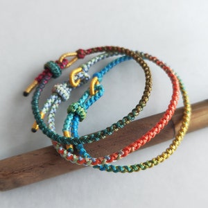 Op de afbeelding: Drie kleurrijke gevlochten armbanden met verschillende kleuren, waaronder rood, oranje, geel, groen, blauw en wit. De armbanden zijn gemaakt van dun koord en zijn aan elkaar geknoopt.