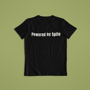 以下が含まれることがあります： 胸に白い文字で「Powered by Spite」と書かれた黒いTシャツ。クルーネックのTシャツで、綿または同様の素材で作られている可能性があります。背景は無地のオリーブグリーンです。