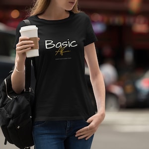 Peut inclure: Un t-shirt noir avec le texte "Basic AF" en lettres blanches et dorées. Le t-shirt est porté par une personne tenant une tasse de café blanche.