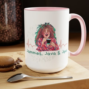 Può includere: Tazza in ceramica bianca con bordo e manico rosa. La tazza presenta un'illustrazione colorata di una donna con i capelli lunghi, che indossa una camicia a quadri rossi e bianchi, tenendo una tazza di caffè. Il testo "Jammies, Java & Jovi" è stampato sulla tazza in verde.