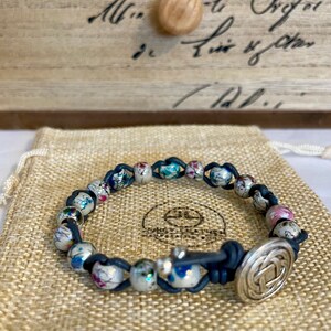 May include: A white and blue beaded bracelet with a silver clasp. The beads are small and have a floral pattern. The bracelet is on a burlap background.