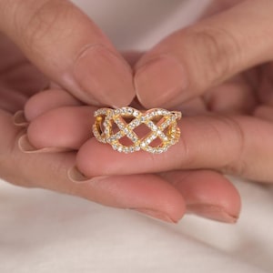 May include: A gold ring featuring a lattice design, set with small, clear gemstones. The ring's intricate pattern is highlighted as it is held between two hands. This piece of jewellery has a sparkling appearance.