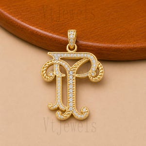 May include: A gold-colored pendant in the shape of the letter "T", encrusted with small, clear gemstones. The pendant has an ornate, scrolled design and a loop at the top for a chain. The background is a warm, neutral tone.