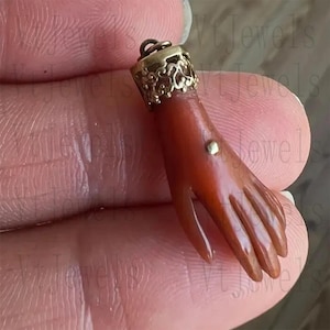 May include: A carved coral pendant in the shape of a hand, featuring a gold-colored filigree cap and a small gold accent. The pendant is a reddish-orange color and has detailed finger and palm features.