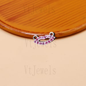 May include: A small, silver-toned charm in the shape of lips, encrusted with pink gemstones. The charm has two small loops for attachment. The background is a warm, neutral color.