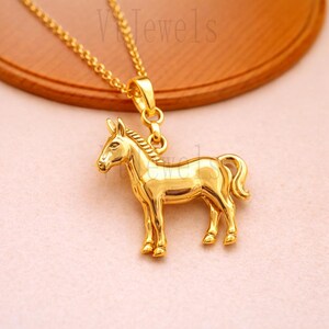 Op de afbeelding: Een goudkleurige ketting met een gedetailleerde paardenhanger. Het paard staat met opgeheven hoofd. De ketting is ook goudkleurig. De tekst "V Jewels" is zichtbaar op de achtergrond.