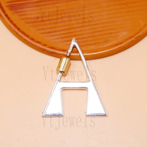 Pode incluir: Pingente prateado em forma da letra "A" com um componente cilíndrico dourado. O pingente é exibido em uma superfície marrom clara. A joia tem um design moderno e minimalista.