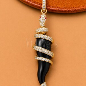 May include: A gold pendant featuring a black horn-shaped stone wrapped in a coiled snake design. The snake is encrusted with small white stones and has two red stones for eyes. The pendant includes a small bail at the top with the text VtJewels.