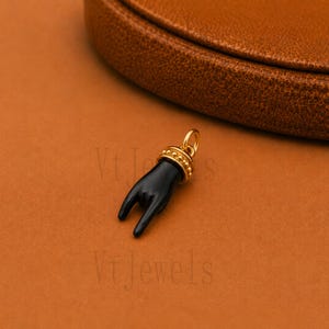 May include: A black hand pendant with a gold-colored ring and accents. The pendant is set against a brown background. The hand is in a gesture with the index and pinky fingers extended. The pendant is likely a charm or jewelry piece.